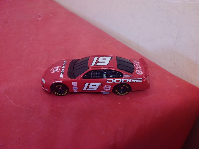 May include: A red Dodge NASCAR diecast car with the number 19 on the side. The car has black accents and Goodyear tires. The car is a replica of the National Training Center car.