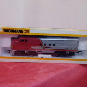 Puede incluir: Una locomotora de tren modelo roja y plateada con las palabras "Santa Fe" impresas en el lateral. La locomotora está en una caja con las palabras "Bachmann HO" impresas en la parte delantera.