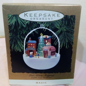 May include: A Keepsake Ornament in a box. The ornament features a miniature Christmas village scene with buildings, a Christmas tree, and figures. The box has text that reads "Keepsake Ornament" and "Last Minute Shopping, Light and Motion!"