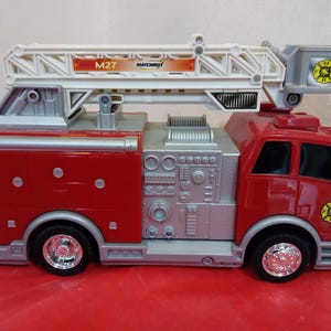 May include: A red and silver toy fire truck with a white extending ladder. The toy truck has black wheels with silver rims and the text "M27 MATCHBOX". The fire truck has a fire department emblem on the side.