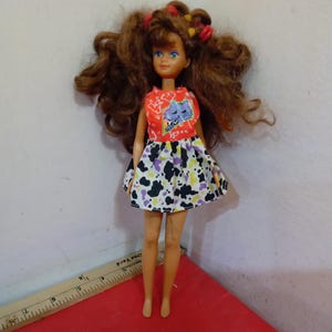 May include: A vintage doll with brown curly hair styled with red and yellow barrettes. The doll wears a red top with a blue cat graphic and a black, white, and purple patterned skirt. The doll is standing on a red surface.