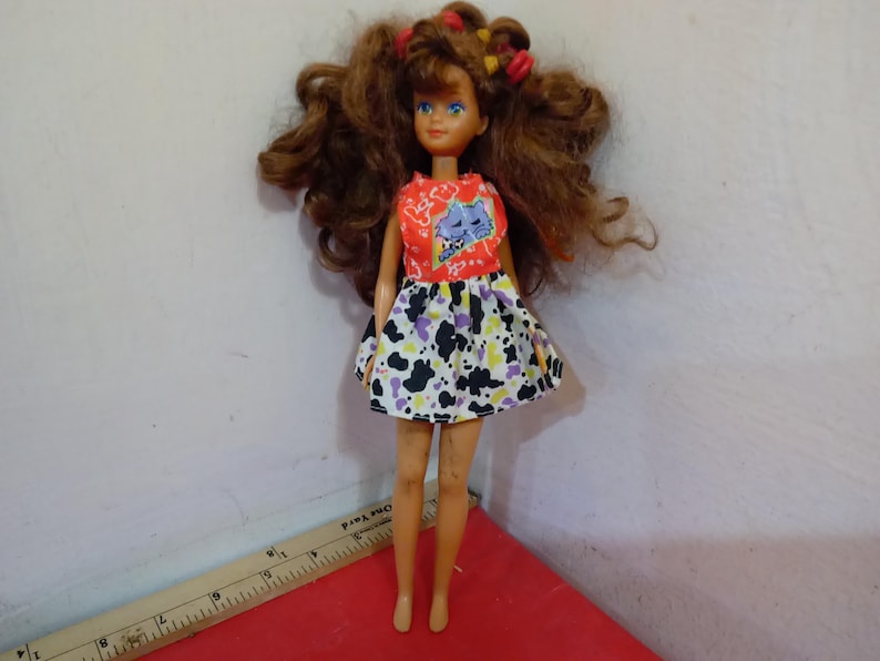 May include: A vintage doll with brown curly hair styled with red and yellow barrettes. The doll wears a red top with a blue cat graphic and a black, white, and purple patterned skirt. The doll is standing on a red surface.