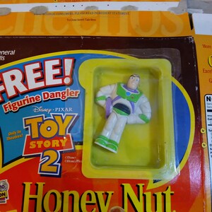 Puede incluir: Una caja de cereal Honey Nut Cheerios de General Mills con un colgador de figura de Toy Story 2 gratis que presenta a Buzz Lightyear. La caja es amarilla y roja con una imagen de Buzz Lightyear con un traje de ranger espacial. La caja tambi&eacute;n incluye una etiqueta de informaci&oacute;n nutricional.