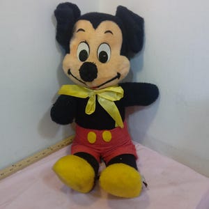 May include: A vintage Mickey Mouse plush toy. The plush features a black body, red shorts with yellow buttons, and yellow shoes. Mickey has a cream-colored face, black ears, and a yellow bow tie. The toy is sitting on a pink surface.