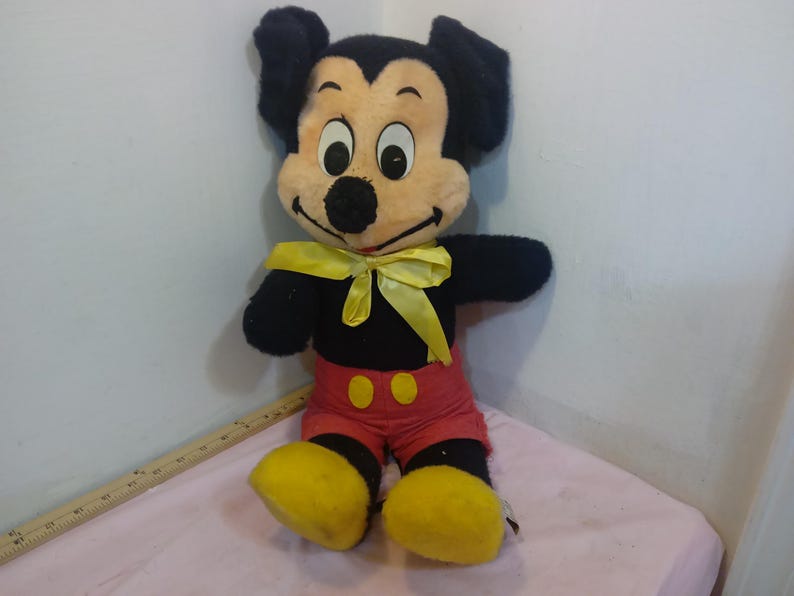 May include: A vintage Mickey Mouse plush toy. The plush features a black body, red shorts with yellow buttons, and yellow shoes. Mickey has a cream-colored face, black ears, and a yellow bow tie. The toy is sitting on a pink surface.
