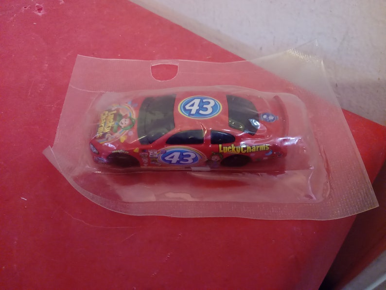 May include: A red toy race car with the number 43 on the side and the words "Lucky Charms" printed on the car. The car is in a clear plastic bag.