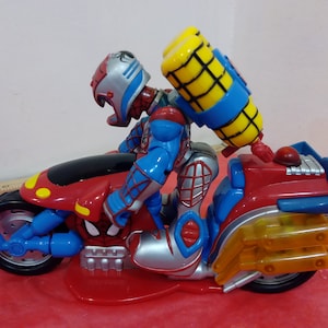 May include: A red, blue, and silver toy motorcycle with a Spider-Man figure riding it. The figure is wearing a red, blue, and silver helmet and has a yellow and blue jetpack on its back.