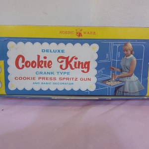 Puede incluir: Una caja azul y amarilla con el texto "Deluxe Cookie King Crank Type Cookie Press Spritz Gun and Basic Decorator" en la parte delantera. La caja muestra a una mujer usando una prensa de galletas y una bandeja de galletas.