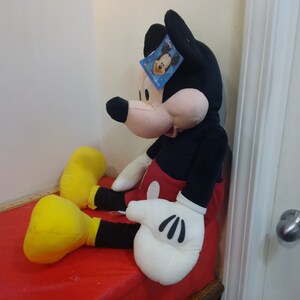 May include: Large plush Mickey Mouse toy sitting on a red surface. Mickey is wearing his signature red shorts with a yellow button and black shoes. He has a black and white face with a big smile and a tag on his ear that says "Disney".
