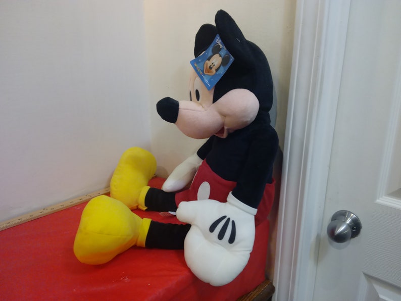 May include: Large plush Mickey Mouse toy sitting on a red surface. Mickey is wearing his signature red shorts with a yellow button and black shoes. He has a black and white face with a big smile and a tag on his ear that says "Disney".