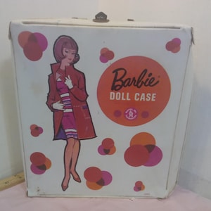 May include: A vintage Barbie doll case with a white exterior. The case features a graphic of a woman in a red coat and the words "Barbie DOLL CASE" in an orange circle. Pink and orange abstract shapes decorate the case.