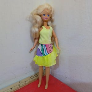 May include: A vintage doll with blonde hair and blue eyes, wearing a yellow tank top and a skirt with a colorful abstract design. The skirt has a yellow ruffle at the bottom. The doll is standing on a red surface.