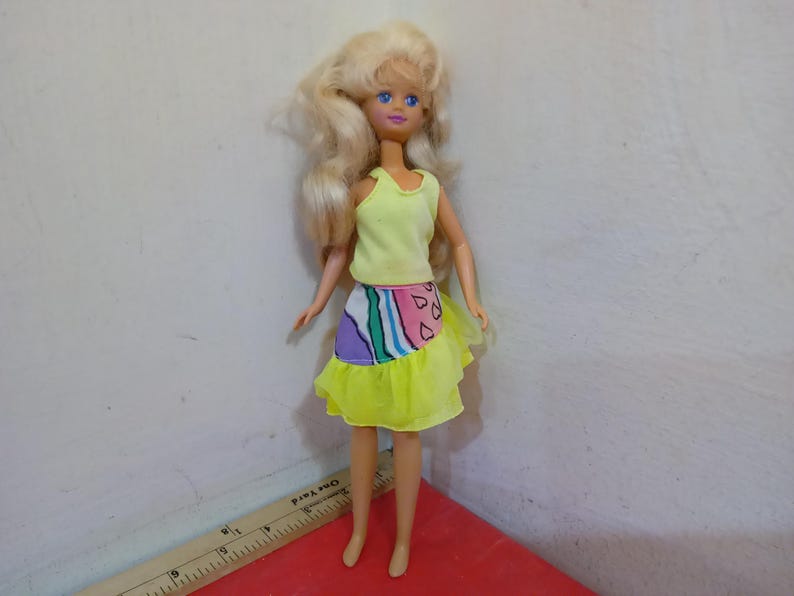 May include: A vintage doll with blonde hair and blue eyes, wearing a yellow tank top and a skirt with a colorful abstract design. The skirt has a yellow ruffle at the bottom. The doll is standing on a red surface.