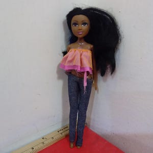 May include: A doll with dark skin, long black hair, and brown eyes. The doll is wearing a pink and orange top and dark blue jeans. The doll is standing on a red surface.