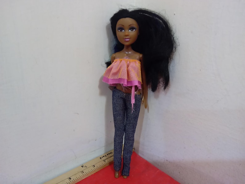 May include: A doll with dark skin, long black hair, and brown eyes. The doll is wearing a pink and orange top and dark blue jeans. The doll is standing on a red surface.