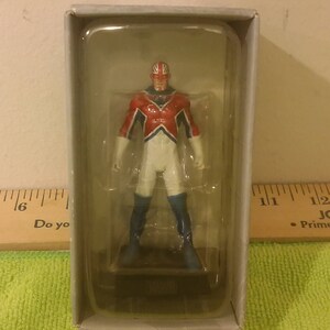 May include: A figurine of a superhero in a clear plastic case. The figure is wearing a red, white, and blue costume with a Union Jack design on the head. The figure is standing on a black base with the word "MARVEL" on it.