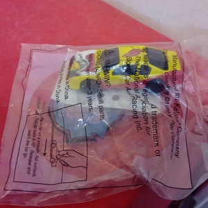 May include: A yellow and red toy race car with a black and white NASCAR logo. The packaging includes a warning about small parts and instructions on how to play with the car. The text on the packaging reads: "Manufactured for Kellogg Company. TM Kellogg Company &copy;1990. NASCAR is a registered trademark of The National Association for Stock Car Auto Racing Inc."