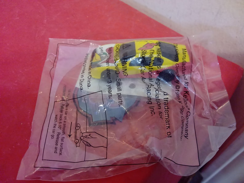 May include: A yellow and red toy race car with a black and white NASCAR logo. The packaging includes a warning about small parts and instructions on how to play with the car. The text on the packaging reads: "Manufactured for Kellogg Company. TM Kellogg Company &copy;1990. NASCAR is a registered trademark of The National Association for Stock Car Auto Racing Inc."