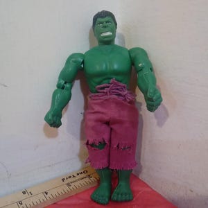 May include: Green plastic action figure of the Hulk wearing a pink, ripped, cloth pants. The figure has a green face with a serious expression and is posed with its arms raised.