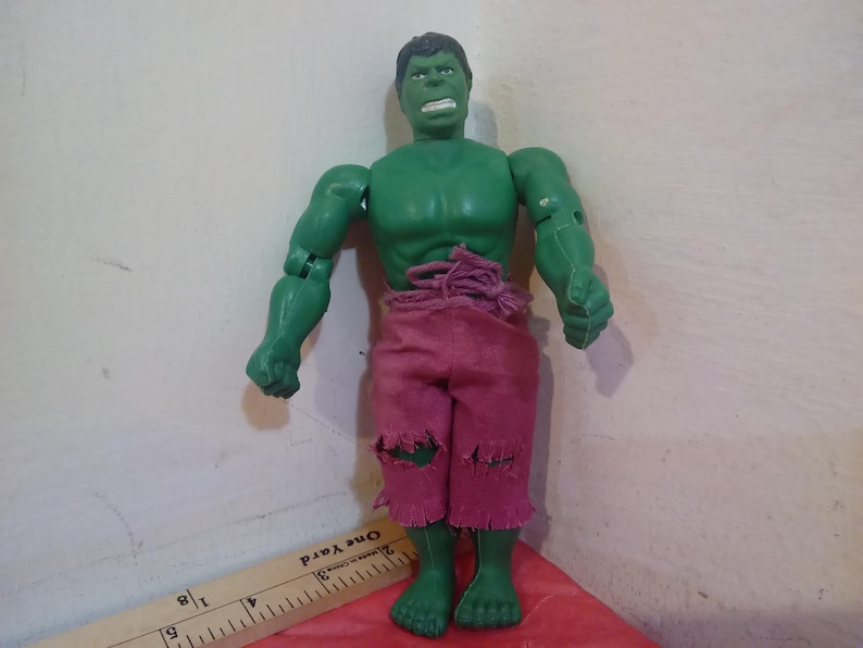 May include: Green plastic action figure of the Hulk wearing a pink, ripped, cloth pants. The figure has a green face with a serious expression and is posed with its arms raised.
