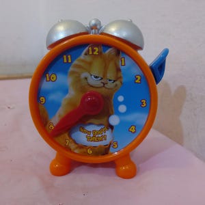 以下が含まれることがあります： オレンジ色のプラスチック製の目覚まし時計で、文字盤に漫画のキャラクター、ガーフィールドの猫が描かれています。文字盤には黒い数字と赤い針があります。文字盤には「Now you're talkin!」という文字が印刷されています。