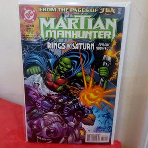 Puede incluir: Portada de cómic de Martian Manhunter, con el personaje principal con un traje verde y rojo y una capa azul. La portada incluye el texto "Rings of Saturn" y "Episode Two of Four."
