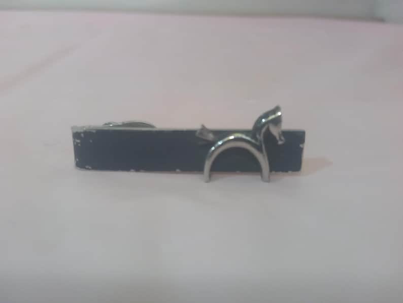 May include: A silver tie clip with a horse design. The clip is attached to a black bar.