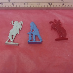 May include: Three plastic toy figures: a light green horse rearing up, a blue Native American figure, and a red cowboy on a bucking bronco. The figures are on a red surface, with a wooden ruler in the background.