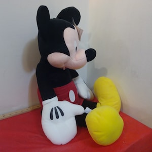 May include: A large plush Mickey Mouse toy sitting on a red surface. Mickey is wearing his signature red shorts with a yellow button and white gloves. He has black ears and a black nose.