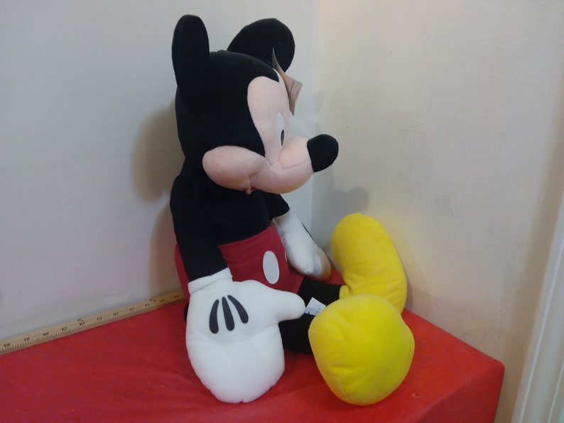 May include: A large plush Mickey Mouse toy sitting on a red surface. Mickey is wearing his signature red shorts with a yellow button and white gloves. He has black ears and a black nose.