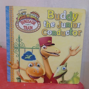 May include: A book cover featuring Buddy the Junior Conductor from Dinosaur Train. The cover is yellow with green, blue, and red accents. The title "Buddy the Junior Conductor" is in large, colorful letters. The book features illustrations of Buddy and other dinosaurs.