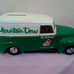May include: A white and green toy delivery van with a Mountain Dew logo and the text "Ya-hoo! It'll tickle your innards." The van has a slot on the roof for coins, suggesting it is a piggy bank.