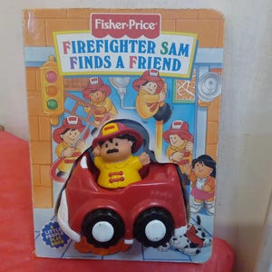 Peut inclure: Un livre de jeu Fisher-Price Little People intitulé "Firefighter Sam Finds a Friend". La couverture présente un camion de pompiers rouge avec une figurine de pompier à l'intérieur. Le livre est illustré avec des personnages colorés.