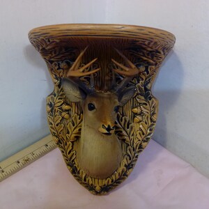 May include: A decorative wall shelf featuring a deer head design. The shelf is made of a light brown material with a detailed relief of a deer head, antlers, and foliage. The color palette includes shades of brown, tan, and cream.