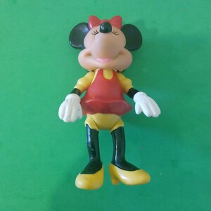 Może przedstawiać: Figurka Myszki Minnie, z czerwoną kokardą, czerwoną sukienką, żółtą górą i żółtymi butami. Figurka ma białe rękawiczki i czarne uszy. Tło jest w jednolitym zielonym kolorze.