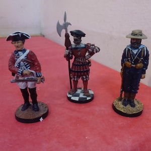 Puede incluir: Tres soldados de juguete en miniatura, cada uno con un uniforme diferente. El soldado de la izquierda lleva un uniforme rojo y blanco con un sombrero. El soldado del centro lleva un uniforme rojo y negro con un sombrero y sostiene una lanza. El soldado de la derecha lleva un uniforme azul con un sombrero y sostiene un rifle.