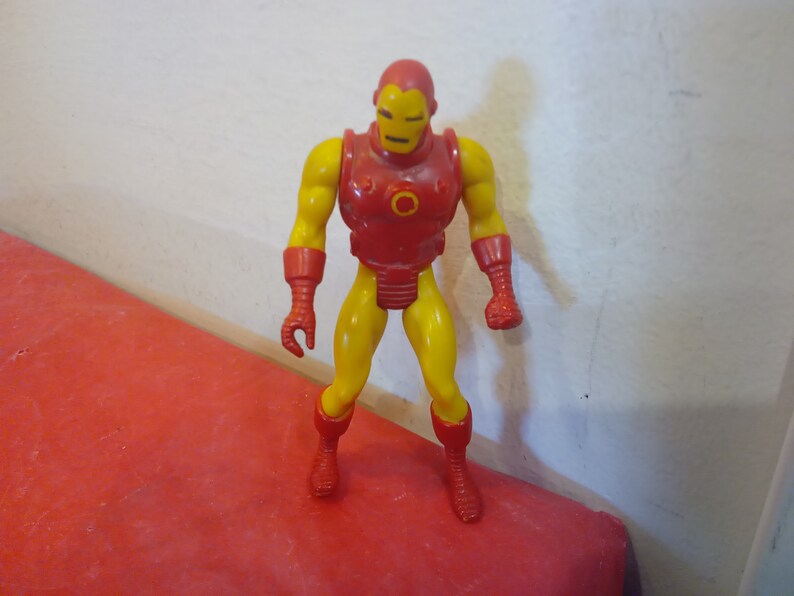 Pode incluir: Uma figura de a&ccedil;&atilde;o vintage do Homem de Ferro, um super-her&oacute;i com um traje vermelho e amarelo, em p&eacute; sobre uma superf&iacute;cie vermelha.