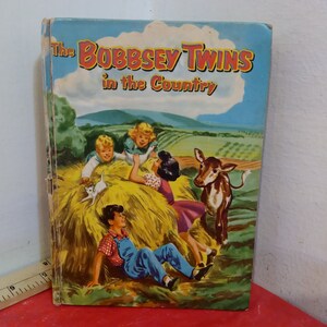 Puede incluir: Una portada de libro vintage con una ilustración de cuatro niños jugando en un campo de heno con un ternero. El título del libro es "The Bobbsey Twins in the Country".