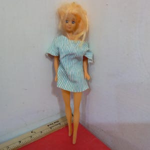 May include: A vintage doll with blonde hair and blue eyes, wearing a short, light blue and white striped dress. The doll is standing on a red surface, with a ruler in the foreground.