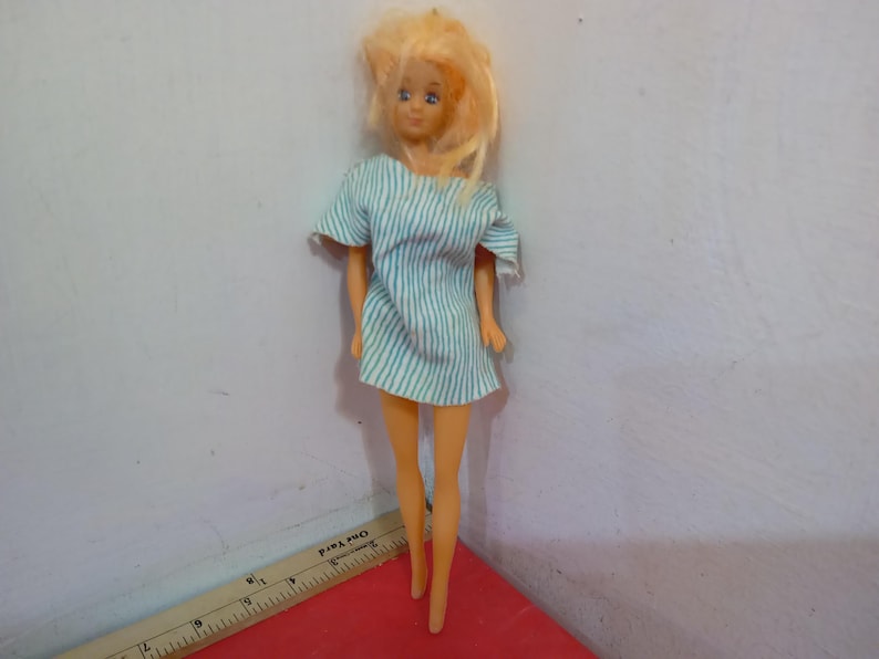 May include: A vintage doll with blonde hair and blue eyes, wearing a short, light blue and white striped dress. The doll is standing on a red surface, with a ruler in the foreground.
