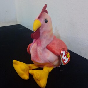 Może przedstawiać: Różowo-czerwony beanie baby koguta z żółtym dziobem i stopami. Beanie baby ma czerwoną metkę Ty z białą gwiazdą.