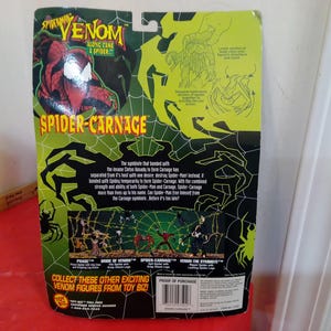 Może przedstawiać: Opakowanie figurki Spider-Man Venom z Spider-Carnage. Opakowanie jest gł&oacute;wnie zielono-czarne z czerwono-czarną grafiką Spider-Carnage. Tekst "Spider-Carnage" jest wyraźnie widoczny.