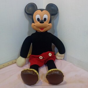 May include: A plush Mickey Mouse doll with a black shirt, red shorts, and brown shoes. The doll has large black ears, a tan face, and white gloves. The doll is sitting on a pink surface.