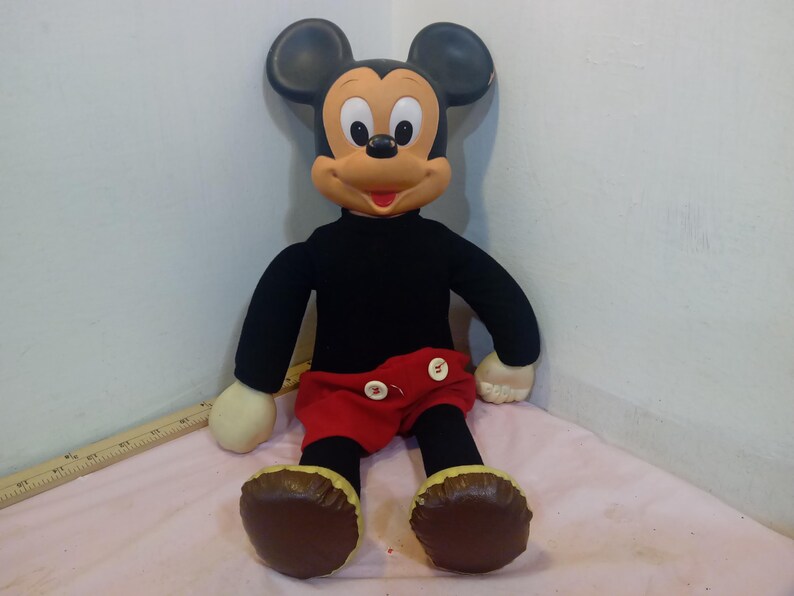 May include: A plush Mickey Mouse doll with a black shirt, red shorts, and brown shoes. The doll has large black ears, a tan face, and white gloves. The doll is sitting on a pink surface.