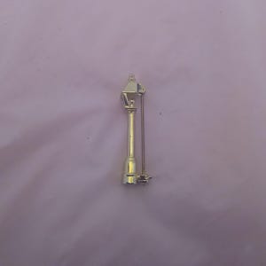 May include: A gold-toned metal brooch in the shape of a street lamp. The lamp has a pointed top and a round base. The brooch has a pin on the back.