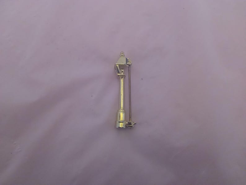 May include: A gold-toned metal brooch in the shape of a street lamp. The lamp has a pointed top and a round base. The brooch has a pin on the back.