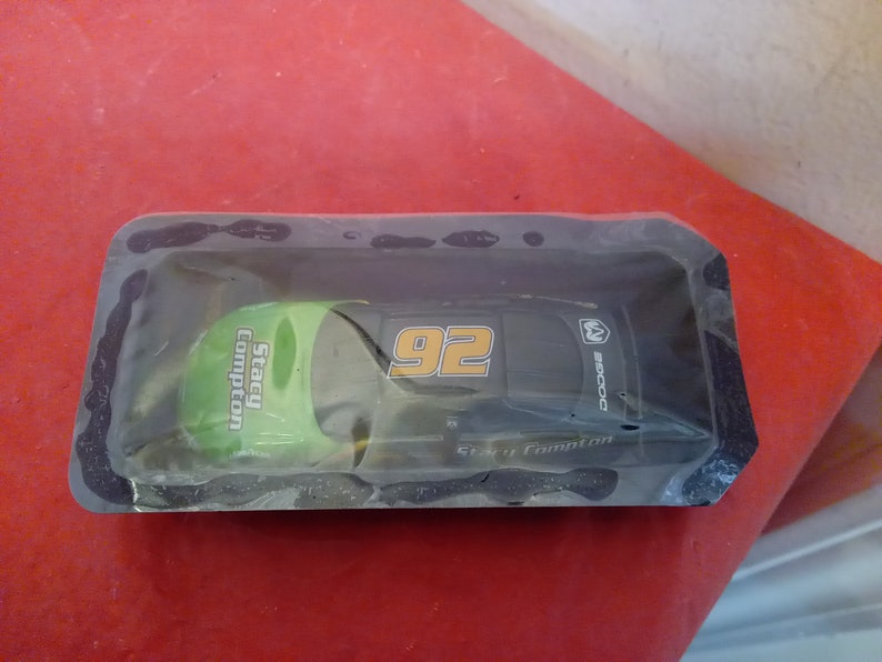 May include: A black and green toy race car with the number 92 and the name Stacy Compton on the side. The car is in a plastic package.