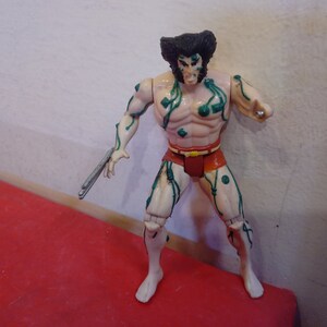 Pode incluir: Uma figura de a&ccedil;&atilde;o de pl&aacute;stico do Wolverine dos X-Men, com videiras verdes crescendo sobre seu corpo. Ele est&aacute; vestindo um pano de renda vermelho e tem uma garra de prata na m&atilde;o.