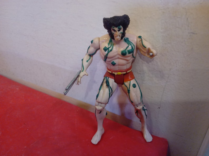 Pode incluir: Uma figura de a&ccedil;&atilde;o de pl&aacute;stico do Wolverine dos X-Men, com videiras verdes crescendo sobre seu corpo. Ele est&aacute; vestindo um pano de renda vermelho e tem uma garra de prata na m&atilde;o.