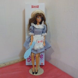 Puede incluir: Una muñeca Little Debbie con un vestido de cuadros azules y blancos, un delantal blanco y un sombrero de paja. La muñeca sostiene una espátula de plástico azul y está de pie sobre un soporte blanco. La muñeca está empaquetada en una caja con el logotipo de Little Debbie.